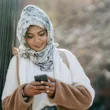 cheerful young ethnic female in headscarf and coat browsing smartphone