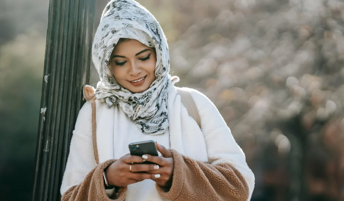 cheerful young ethnic female in headscarf and coat browsing smartphone