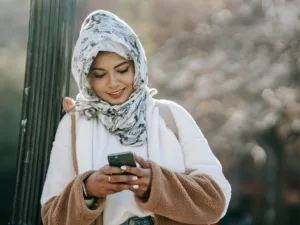 cheerful young ethnic female in headscarf and coat browsing smartphone