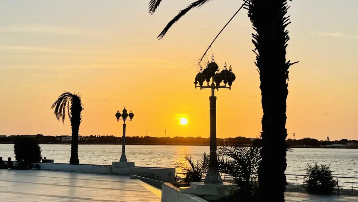 A picturesque sunset over the Jeddah Waterfront promenade. Silhouetted palm trees frame the scene, while elegant streetlamps stand against the vibrant orange and yellow sky.