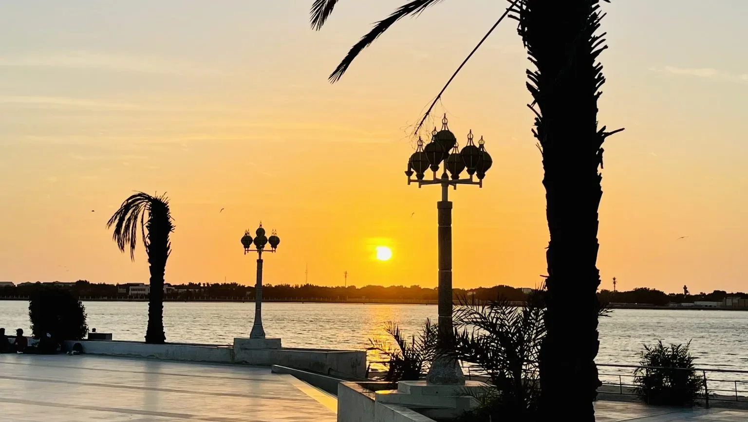 A picturesque sunset over the Jeddah Waterfront promenade. Silhouetted palm trees frame the scene, while elegant streetlamps stand against the vibrant orange and yellow sky.